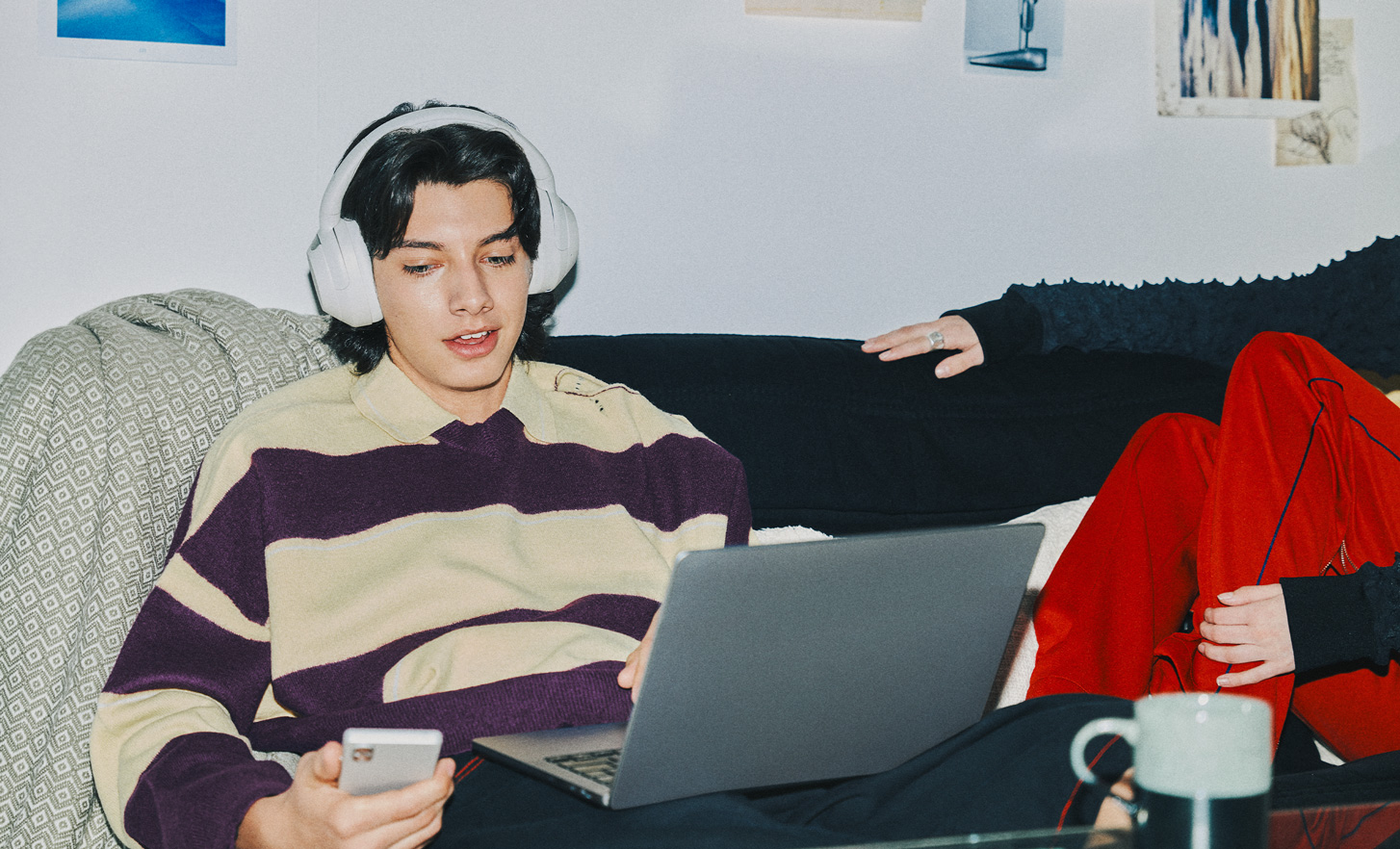 A young person sitting at their laptop with a phone in their hand and wearing the ULT WEAR headphones in white.