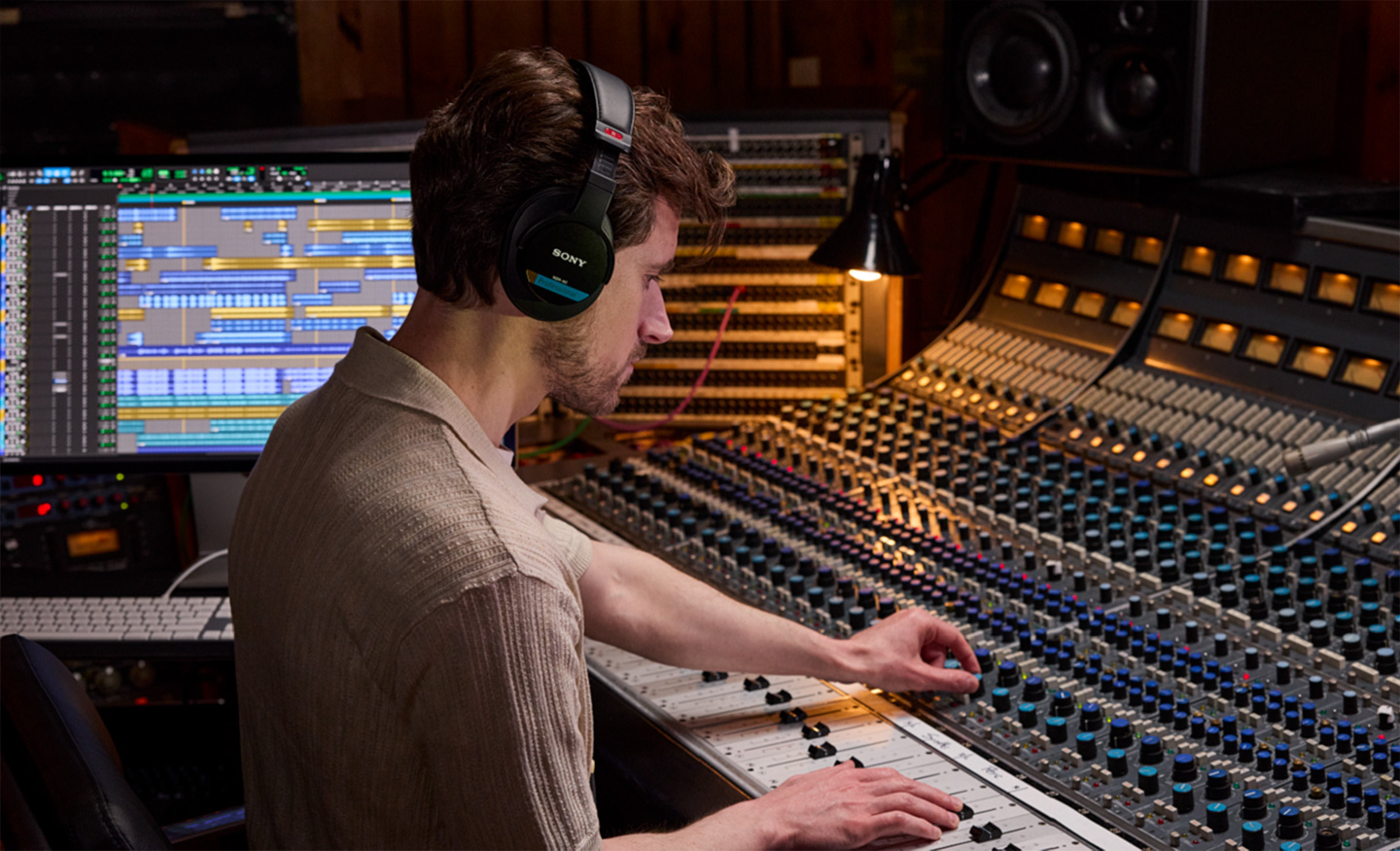  Sound engineer sat down tweaking knobs on his mixing soundboard inside his studio. He is wearing the MDR-M1 Monitor Headphones as he mixes the audio.