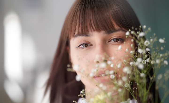 A portrait photograph of a white woman with bangs and flowing hair, facing the camera directly, with a beautiful bokeh effect created by the presence of baby's breath flowers in front of her face.